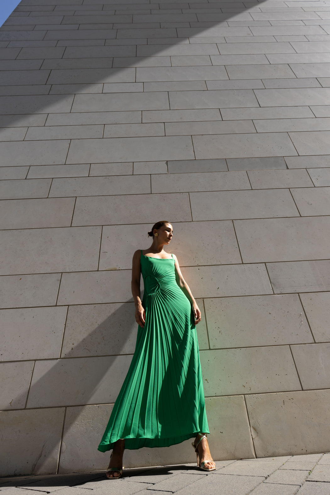 Model in a green dress pleated in contrasting directions standing against a stone wall.