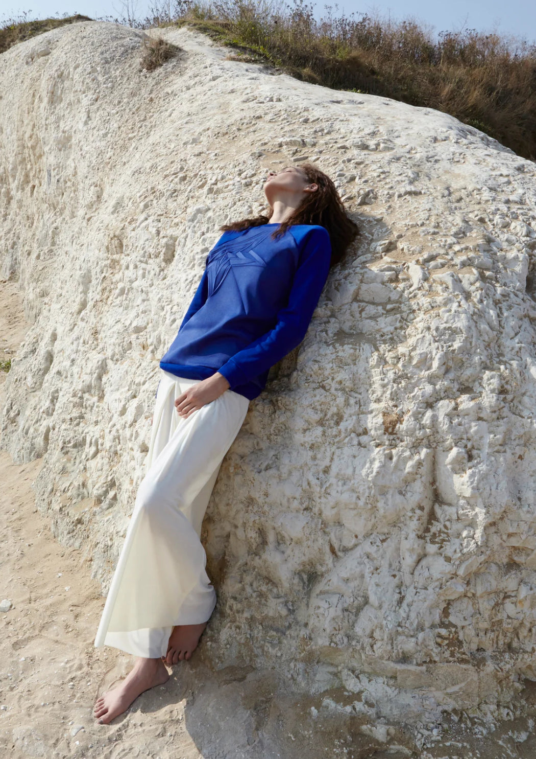A woman wearing a blue jumper and white pants leaning against a large rock.