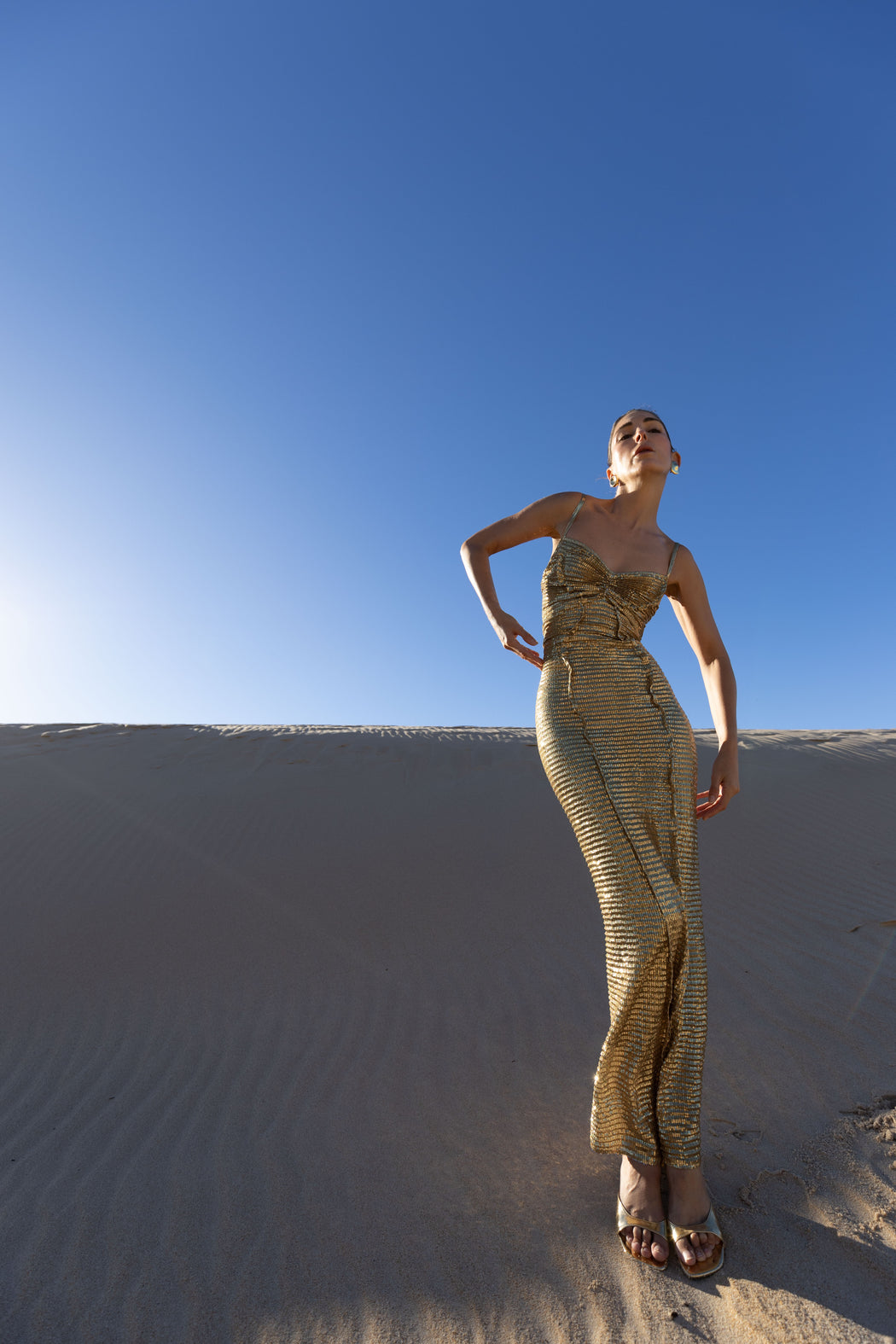 Model in a gold pleated ankle-length sequin dress with unique tuck detailing down the front with a separate bamboo jersey lace standing on a sand dune under a clear blue sky.