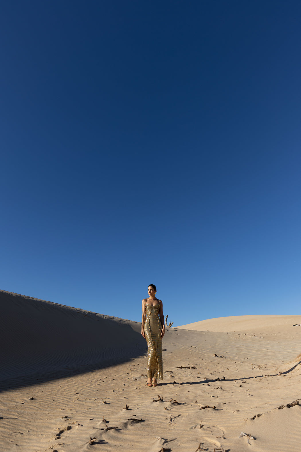 Model in a gold pleated ankle-length sequin dress with unique tuck detailing down the front with a separate bamboo jersey lace standing on a sand dune under a clear blue sky.