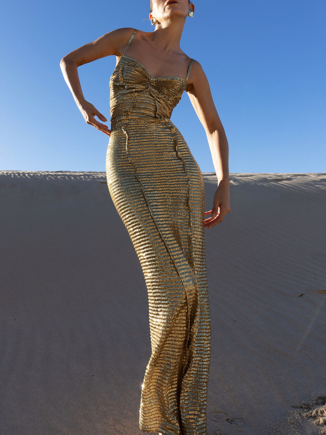Model in a gold pleated ankle-length sequin dress with unique tuck detailing down the front with a separate bamboo jersey lace standing on a sand dune against a clear blue sky.