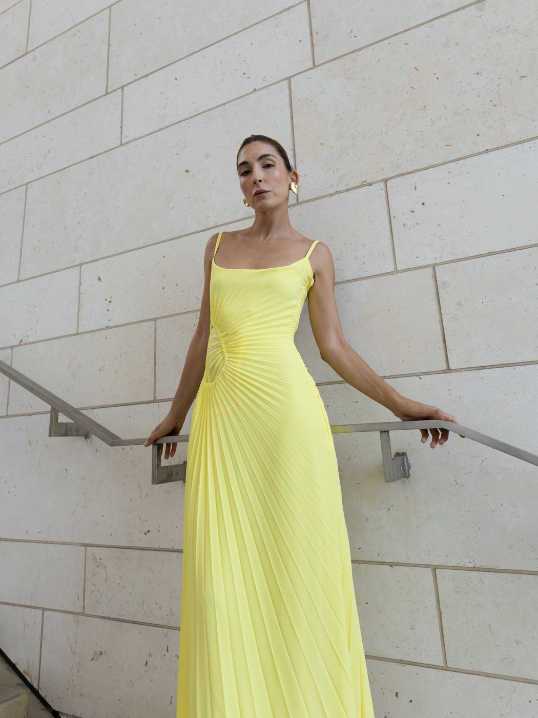 Model in a green georgette dress pleated in contrasting directions standing against a light stone wall.