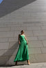 Model in a green dress pleated in contrasting directions standing against a stone wall.