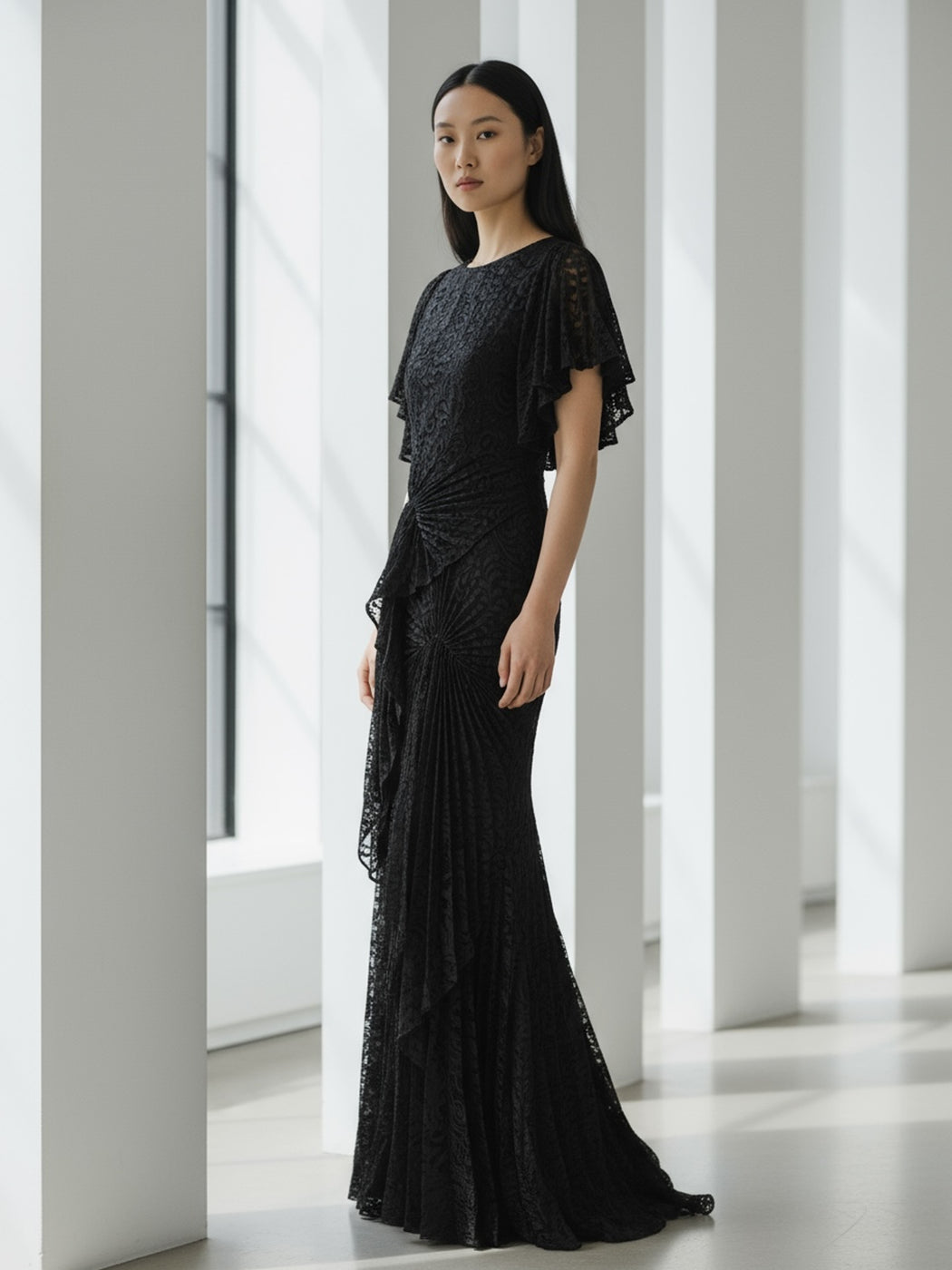 Model wearing a black floor-length dress pleated in a waterfall flowing design with bamboo jersey lining standing in a modern indoor setting