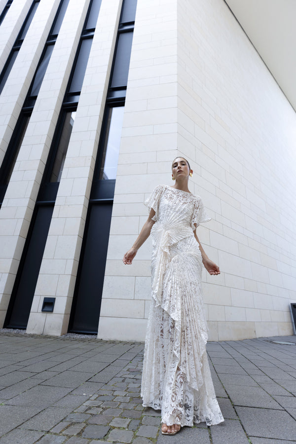 Model in a white floor-length dress pleated in a waterfall flowing design with bamboo jersey lining standing in front of a white modern building.