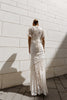 Model in a White floor-length dress pleated in a waterfall flowing design with bamboo jersey lining standing against a light stone wall.