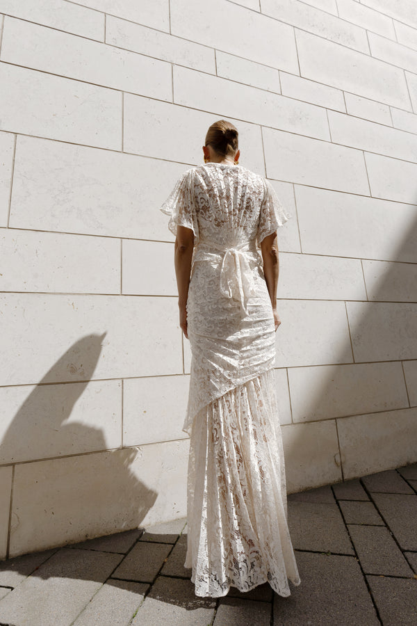 Model in a White floor-length dress pleated in a waterfall flowing design with bamboo jersey lining standing against a light stone wall.
