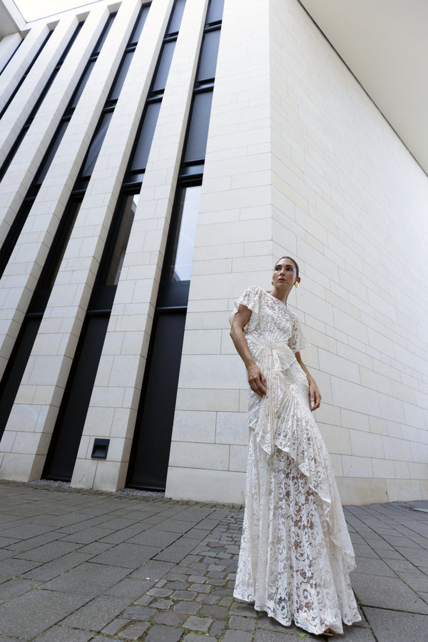 Model in white floor-length dress pleated in a waterfall flowing design with bamboo jersey lining standing against a modern building.