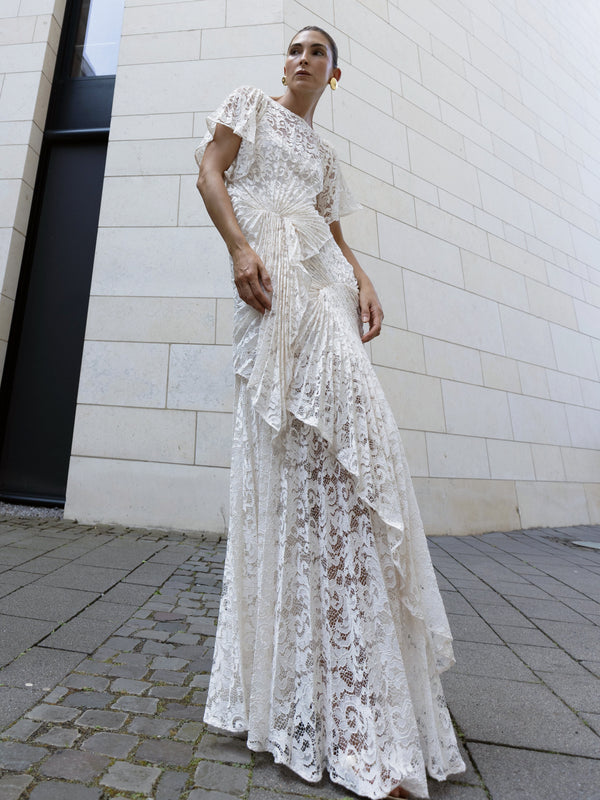 Model wearing a white floor-length dress pleated in a waterfall flowing design with bamboo jersey lining standing against a light-coloured wall.
