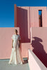 Model wearing a striking sunray design ivory-coloured top, featuring a unique gathered front detail and a draped asymmetric hemline that adds movement and dimension. Crafted from exquisite French Baroque-style lace, it flows gracefully to create a flattering silhouette, standing in front of pink architectural walls with a clear blue sky.