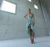 Model in a light blue evening dress with shirred elastic pattern standing in a room with concrete walls and large windows.