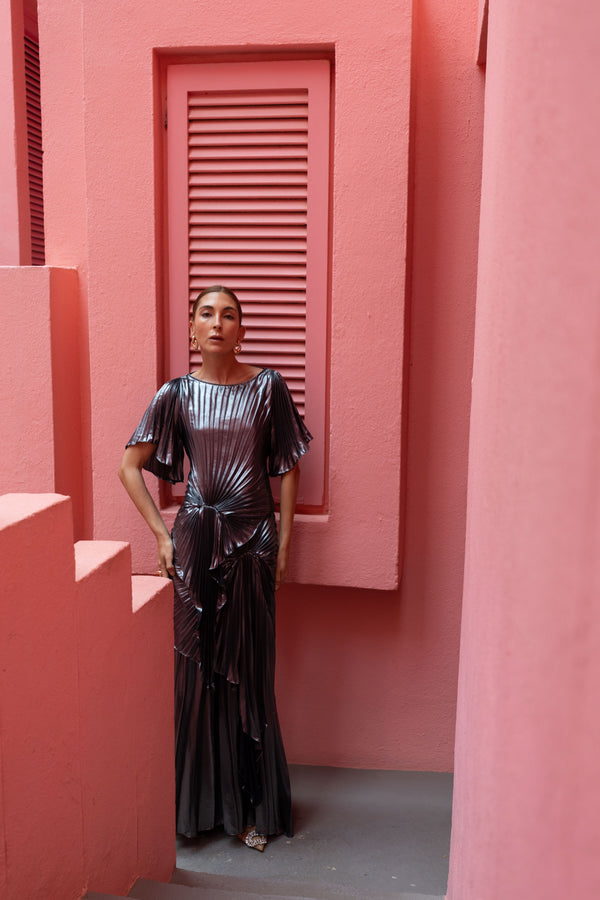 Model wearing a floor length metallic graphite chiffon dress, features delicate pleating in a flowing waterfall design, with a high neckline, slip on style, and adjustable waist tie, standing against a pink wall.