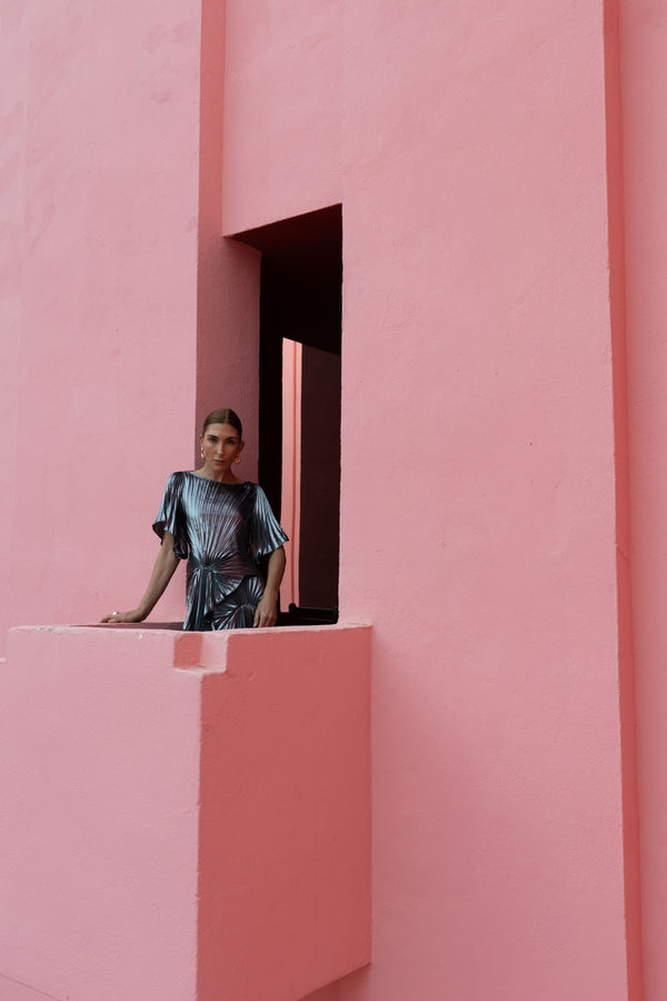 Model wearing a floor length metallic graphite chiffon dress, features delicate pleating in a flowing waterfall design, with a high neckline, slip on style, and adjustable waist tie.