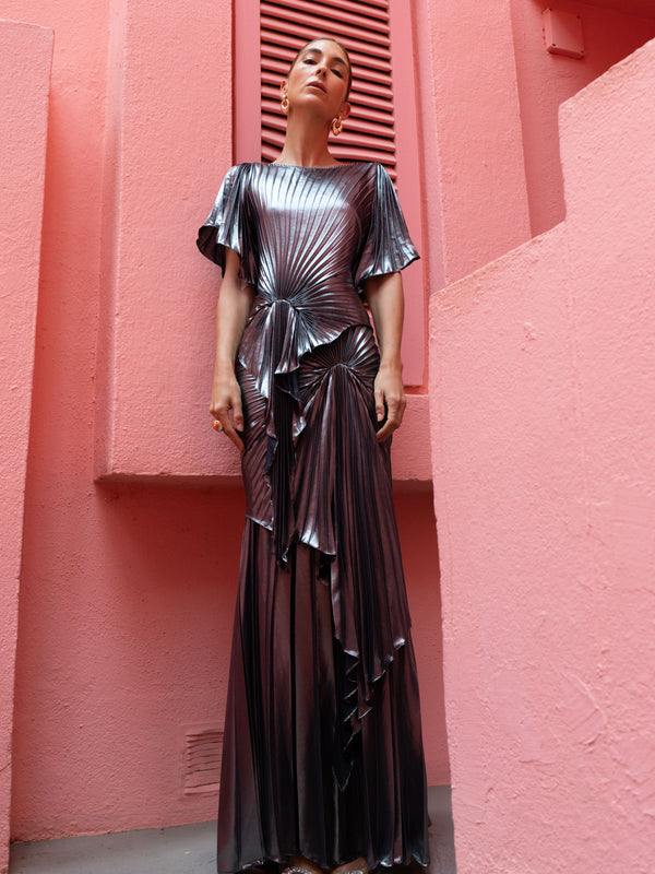 Model wearing a floor length metallic graphite chiffon dress, features delicate pleating in a flowing waterfall design, with a high neckline, slip on style, and adjustable waist tie.