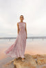 Model wearing a pink sleeveless velvet floor length dress pleated all over in a striking sunray design with side cut out at waist standing on a rock by the river