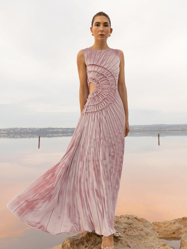 Model wearing a pink sleeveless velvet floor length dress pleated all over in a striking sunray design with side cut out at waist standing on a stone by the river 