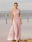Model wearing a pink sleeveless velvet floor length dress pleated all over in a striking sunray design with side cut out at waist standing in a stone by the river