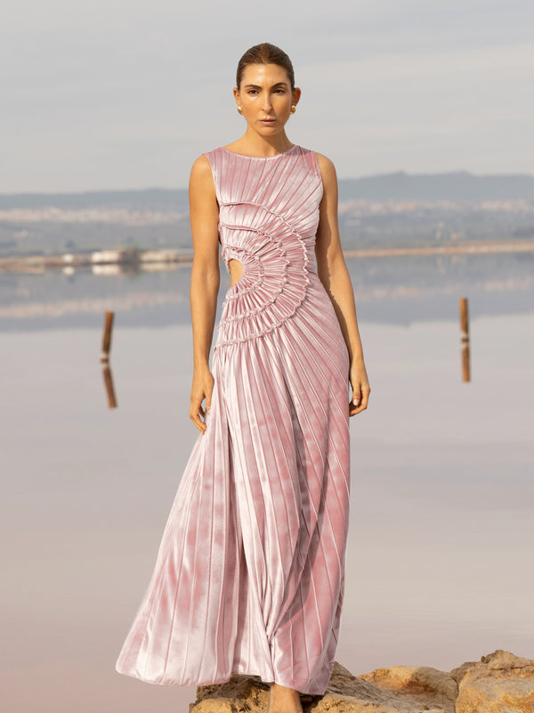 Model wearing a pink sleeveless velvet floor length dress pleated all over in a striking sunray design with side cut out at waist standing in a stone by the river