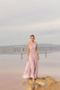 Model wearing a pink sleeveless velvet floor length dress pleated all over in a striking sunray design with side cut out at waist standing on a rock by the river