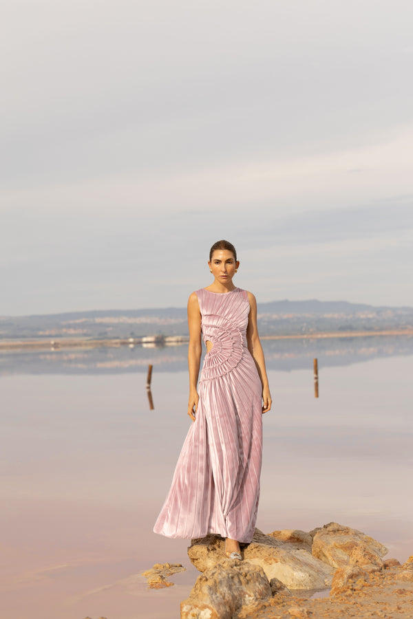 Model wearing a pink sleeveless velvet floor length dress pleated all over in a striking sunray design with side cut out at waist standing on a rock by the river