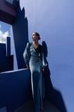 Model wearing a floor length blue velvet wrap dress with puff sleeves, a side waist tie, and an illusion-style sunray pleat draped across the front, creating a fluid, elegant silhouette, standing against a blue wall with architectural elements.