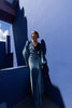 Model wearing a floor length blue velvet wrap dress with puff sleeves, a side waist tie, and an illusion-style sunray pleat draped across the front, creating a fluid, elegant silhouette, standing against a blue wall with architectural elements.