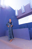 Model wearing a floor length blue velvet wrap dress with puff sleeves, a side waist tie, and an illusion-style sunray pleat draped across the front, creating a fluid, elegant silhouette, standing against a blue wall with architectural elements.