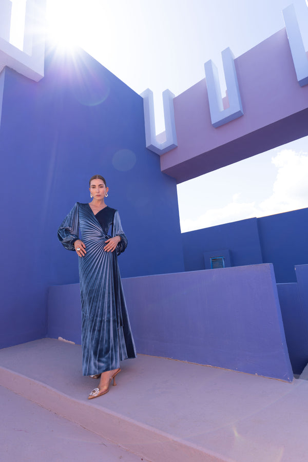 Model wearing a floor length blue velvet wrap dress with puff sleeves, a side waist tie, and an illusion-style sunray pleat draped across the front, creating a fluid, elegant silhouette, standing against a blue wall with architectural elements.