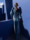Model wearing a floor length velvet wrap dress with puff sleeves, a side waist tie, and an illusion-style sunray pleat draped across the front, creating a fluid, elegant silhouette, standing against a blue wall with architectural elements.