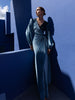Model wearing a floor length velvet wrap dress with puff sleeves, a side waist tie, and an illusion-style sunray pleat draped across the front, creating a fluid, elegant silhouette, standing against a blue wall with architectural elements.