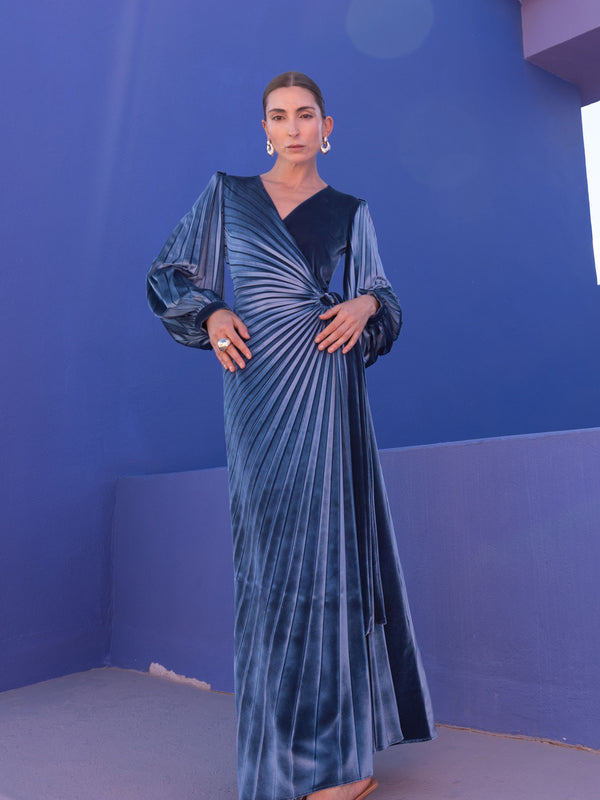 Model wearing a floor-length velvet wrap dress with puff sleeves, a side waist tie, and an illusion-style sunray pleat draped across the front, creating a fluid, elegant silhouette, standing against a blue background.