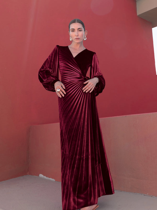 Model wearing a merlot-coloured velvet  wrap style dress pleated in an illusion-style sunray with puff long sleeves standing against a red wall.