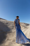 Model wearing a blue metallic sleeveless floor length dress pleated in a sunray pattern with circular shirring design turned away while standing on sand dunes under a clear blue sky