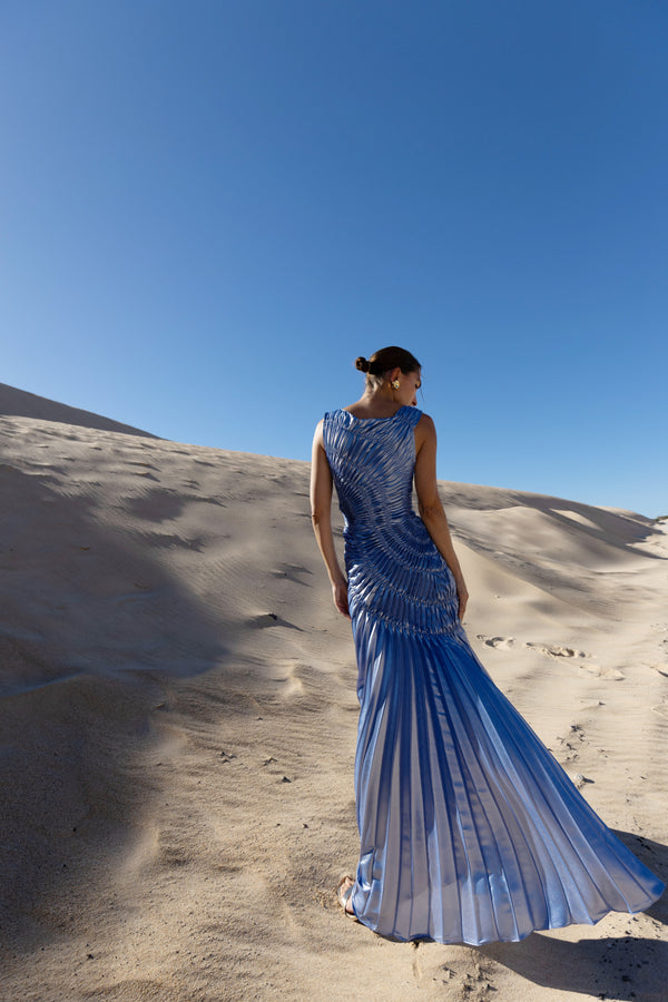 Model wearing a blue metallic sleeveless floor length dress pleated in a sunray pattern with circular shirring design turned away while standing on sand dunes under a clear blue sky