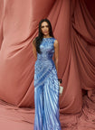 Model wearing a blue metallic sleeveless floor length dress with waist-cinching sunray pleats and elastic contour stitching, standing in front of draped dusty rose fabric.