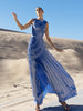 Model wearing a blue metallic sleeveless floor length dress pleated in a sunray pattern with circular shirring design standing on sand dunes with a clear blue sky.