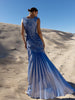 Model wearing a blue metallic sleeveless floor length dress pleated in a sunray pattern with circular shirring design back turned while standing on sand dunes under a clear blue sky.