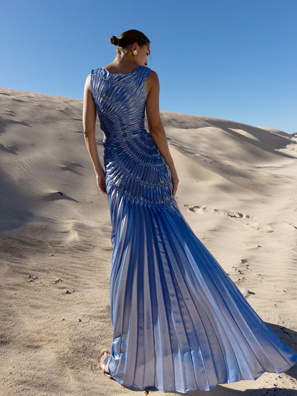 Model wearing a blue metallic sleeveless floor length dress pleated in a sunray pattern with circular shirring design back turned while standing on sand dunes under a clear blue sky.