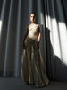 Model wearing an ivory gold metallic floor length dress showcases a unique circular motion design with elastic stitching that contours the body. Pleated in a Sunray pattern, it beautifully flatters the silhouette, standing against a curtain backdrop.