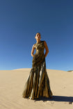 Model wearing a metallic gold sleeveless dress pleated in a sunray pattern with circular shirring design standing on sand dunes with a clear blue sky.