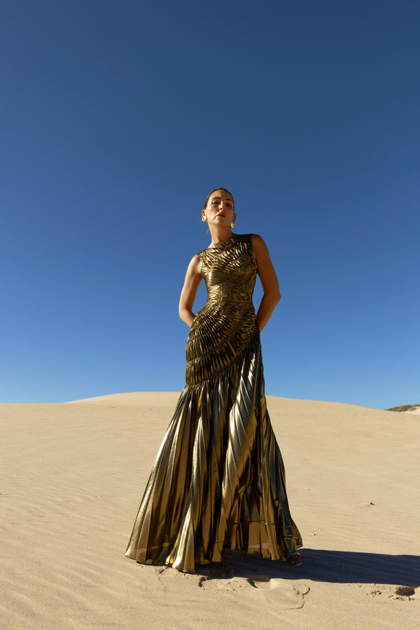 Model wearing a metallic gold sleeveless dress pleated in a sunray pattern with circular shirring design standing on sand dunes with a clear blue sky.