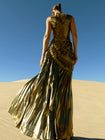 Model wearing a metallic gold sleeveless dress pleated in a sunray pattern with circular shirring design standing on sand dunes with a clear blue sky.