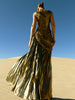 Model wearing a metallic gold sleeveless dress pleated in a sunray pattern with circular shirring design standing on sand dunes with a clear blue sky.