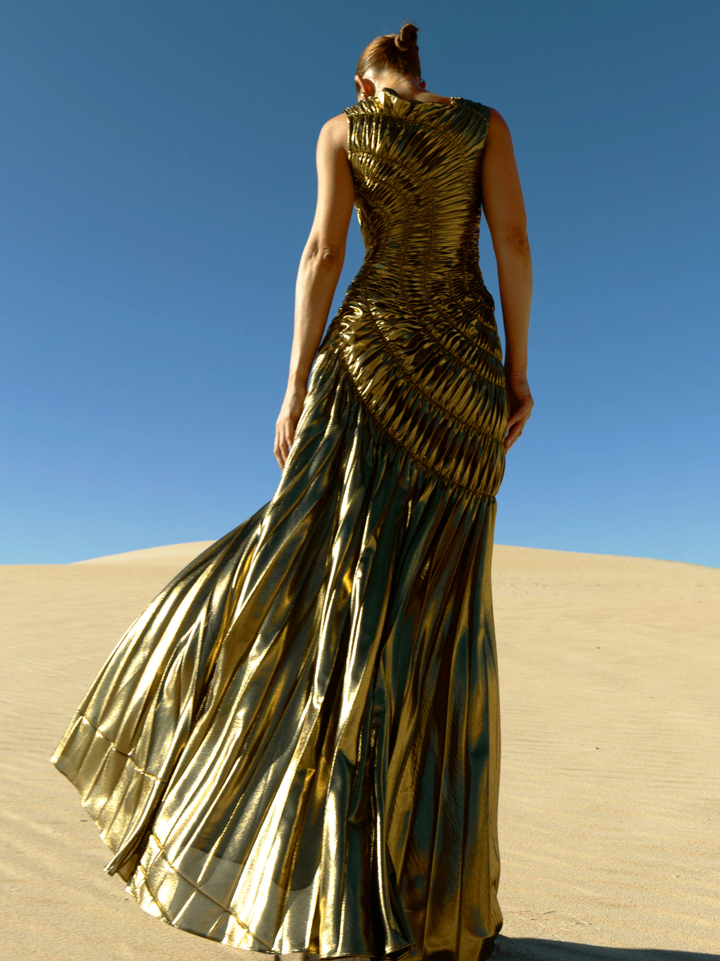 Model wearing a metallic gold sleeveless dress pleated in a sunray pattern with circular shirring design standing on sand dunes with a clear blue sky.