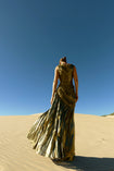 Model wearing a metallic gold sleeveless dress pleated in a sunray pattern with circular shirring design back turned while standing on sand dunes with a clear blue sky.