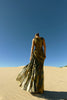 Model wearing a metallic gold sleeveless dress pleated in a sunray pattern with circular shirring design back turned while standing on sand dunes with a clear blue sky.