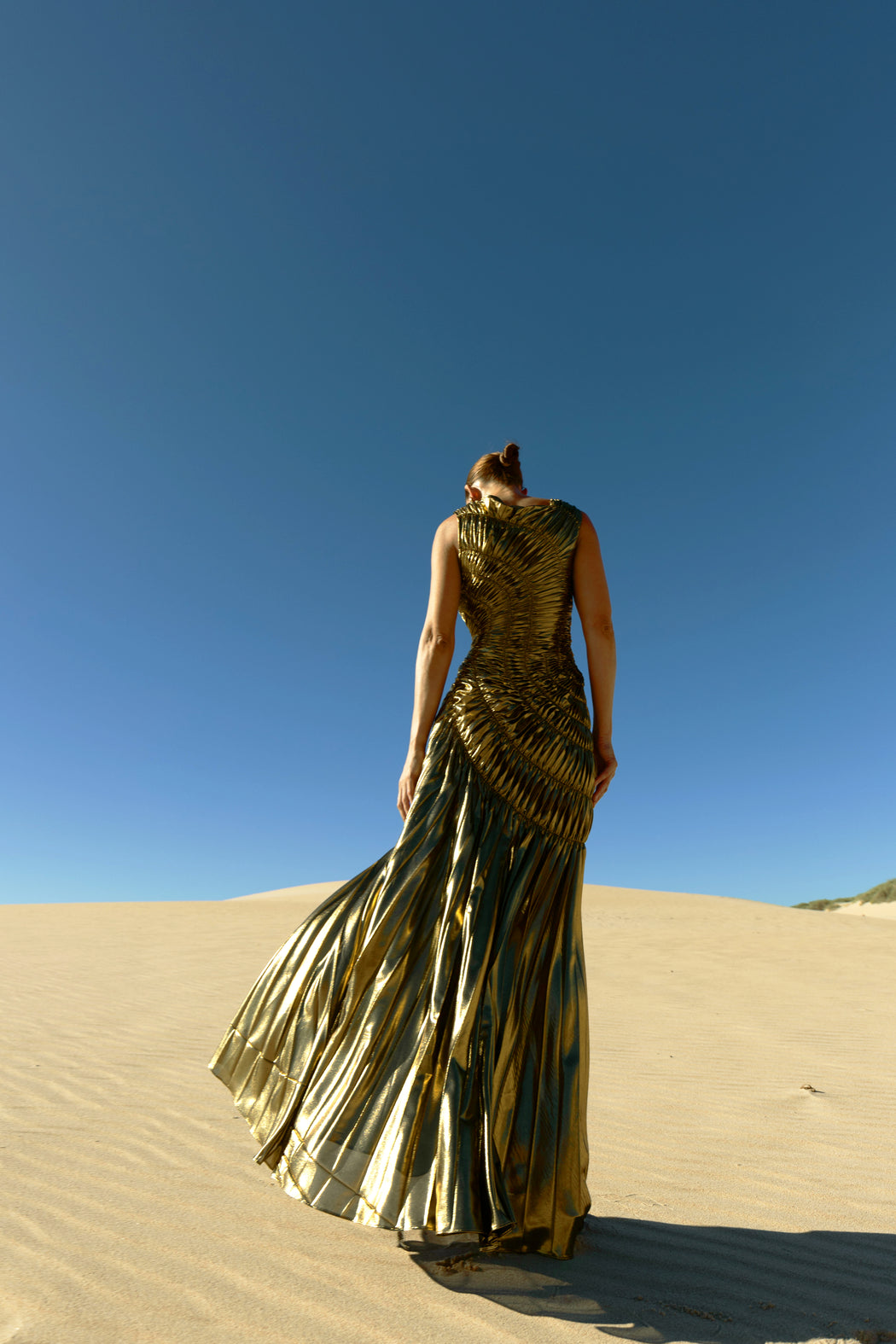 Model wearing a metallic gold sleeveless dress pleated in a sunray pattern with circular shirring design back turned while standing on sand dunes with a clear blue sky.