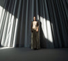 Model  in a metallic gold floor length dress standing in front of a large curtain with dramatic lighting.