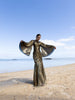 Model wearing a floor length 
metallic pleated dress with a circular elastic shirring design and wide flared sleeves, standing on a beach with a clear blue sky.