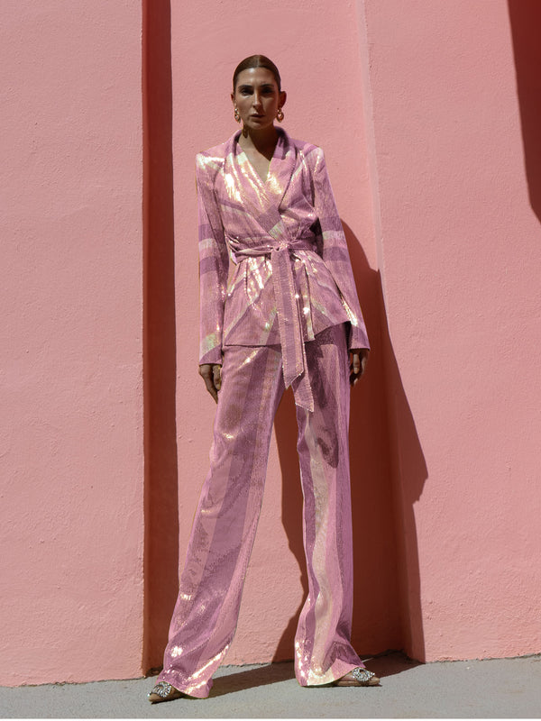 Model wearing a pink wrap-style sequin blazer with sunray stripes and matching wide-leg trousers posing against a pink wall